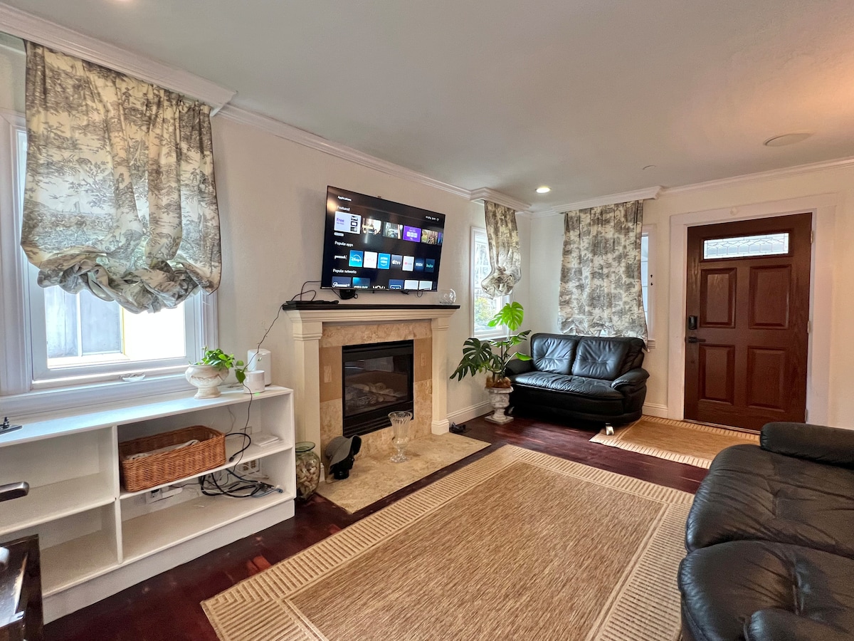 The living room features a comfortable black leather sofa and a second sofa along with a mounted flat-screen TV. Natural light streams through the windows adorned with patterned curtains. A light area rug sits on the dark wooden floor, complementing the warm tones of the space.
