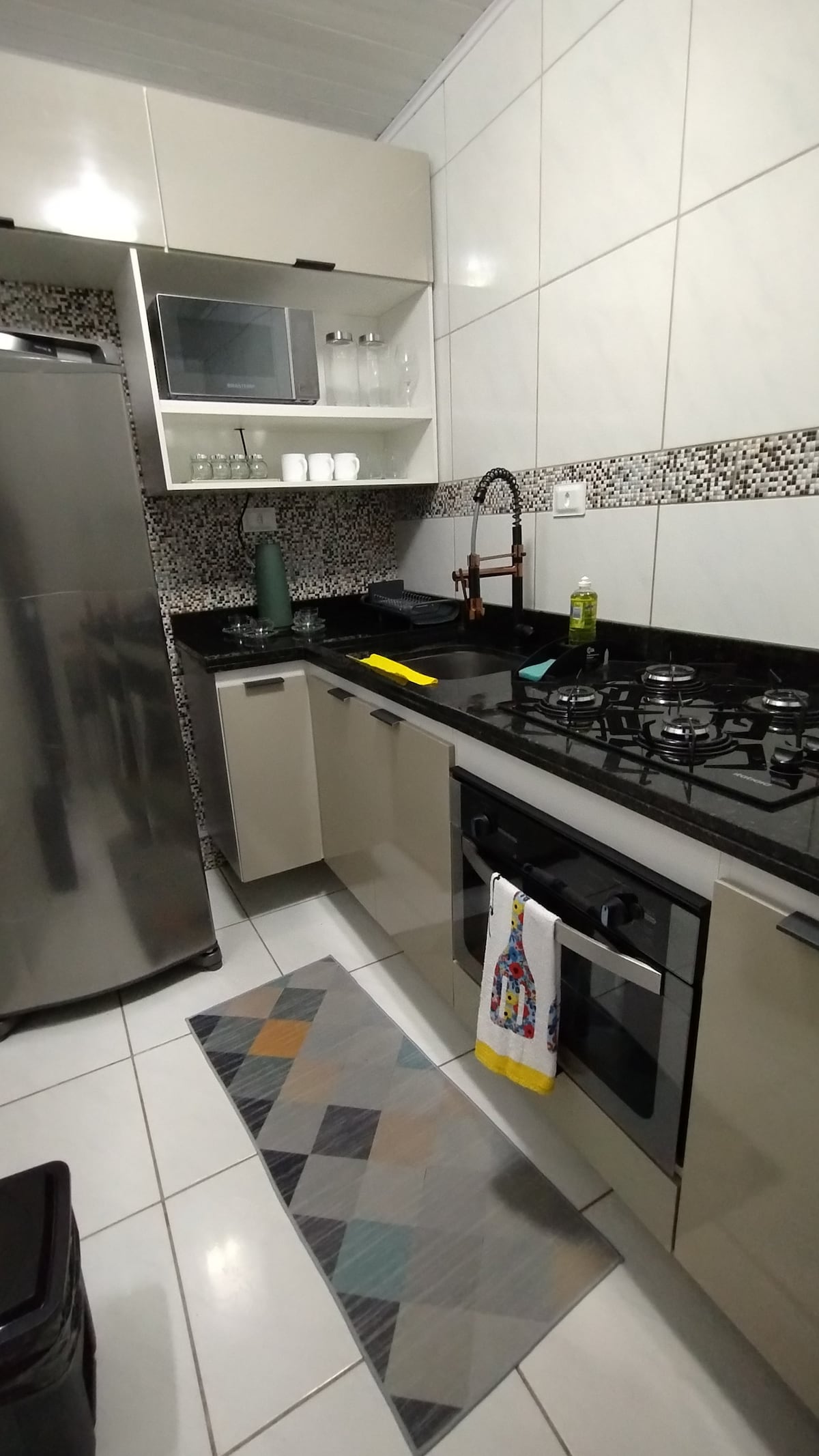 The kitchen features a modern design with sleek cabinets and a dark countertop. Stainless steel appliances, including a refrigerator and a gas stove, are visible. A small rug with a geometric pattern adds color to the floor. Shelves hold glassware and utensils.