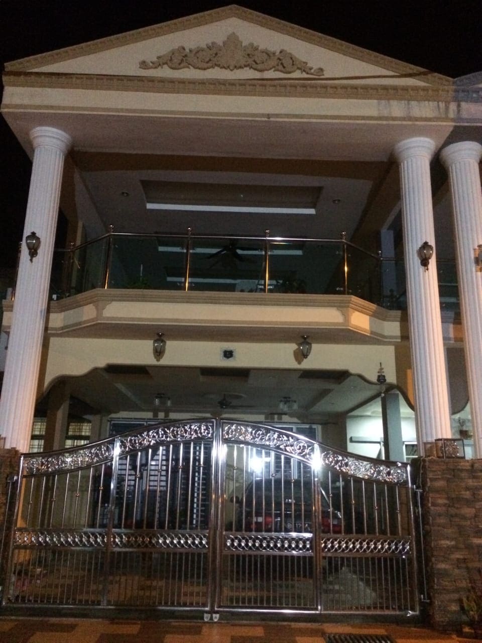 The exterior of the house is captured at night, showing grand columns and a decorative pediment above the entrance. The metallic auto gate adds an elegant touch, while illuminated windows suggest a welcoming interior. The well-lit entrance highlights the spacious facade.