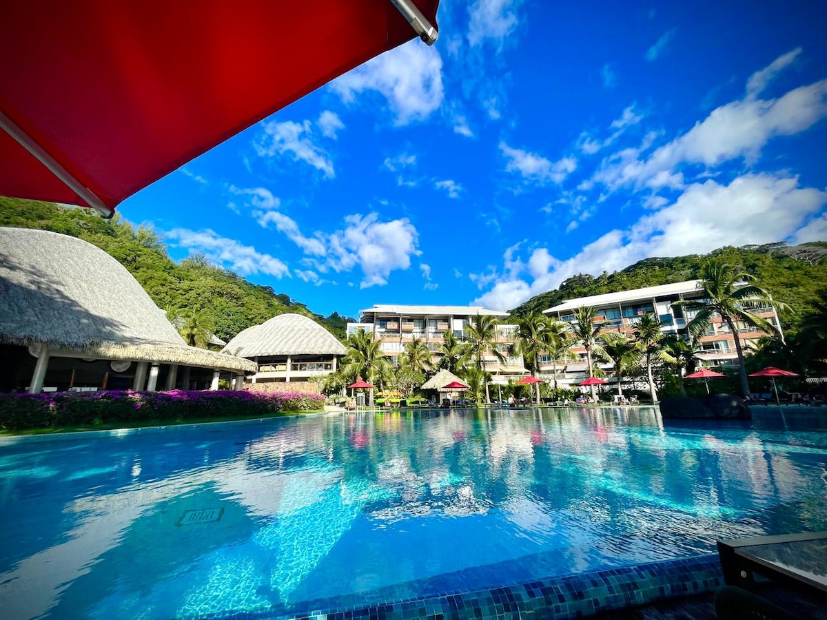 The image depicts a large, inviting swimming pool surrounded by lush greenery and palm trees. The clear blue sky is dotted with fluffy white clouds, while vibrant red umbrellas provide shade for guests. The hotel complex features a thatched-roof structure and multiple levels.