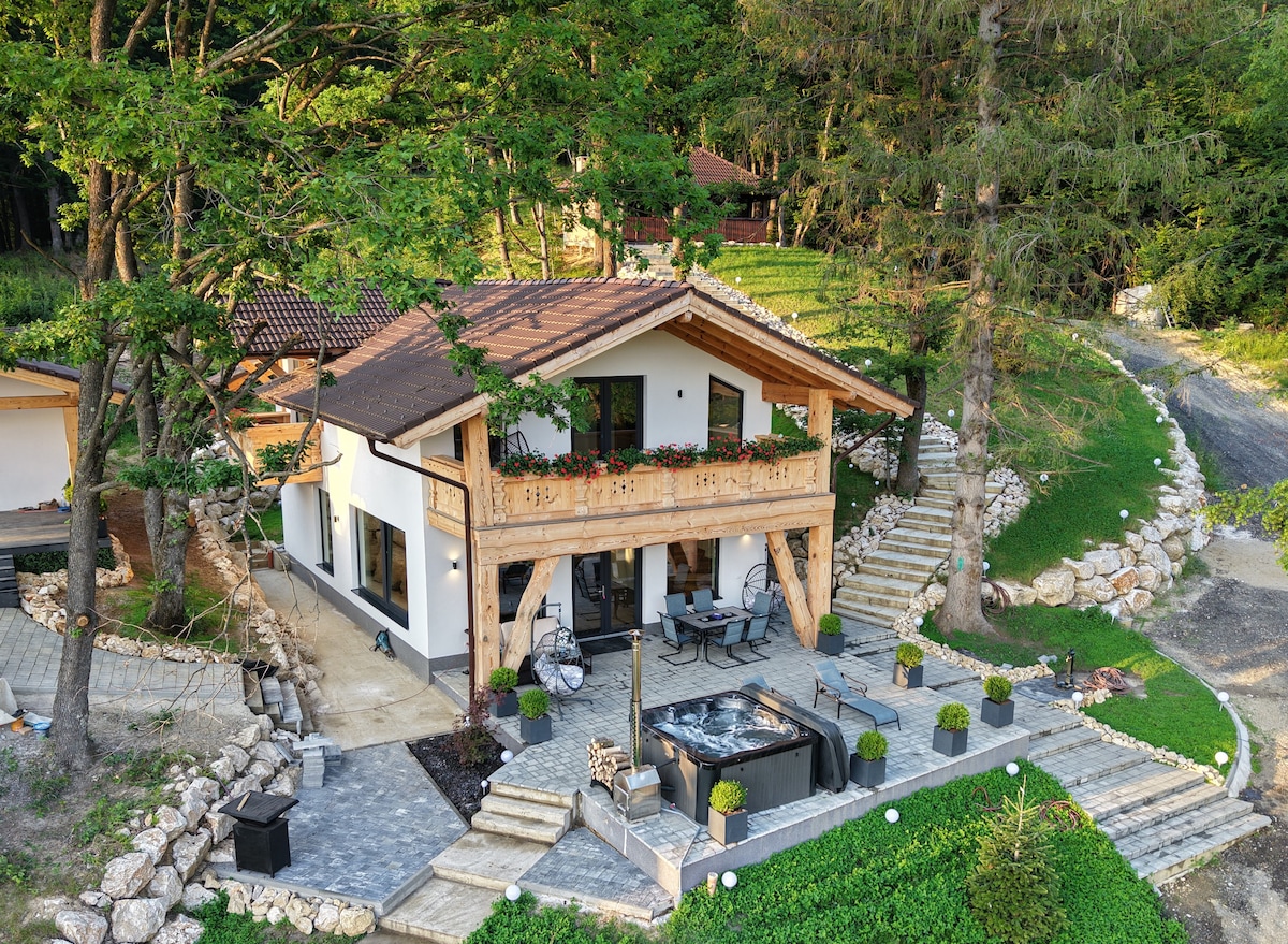 The cabin features a two-story design surrounded by lush greenery, with a terrace adorned with seating arrangements. A hot tub is positioned nearby, while a stone pathway leads to the entrance, highlighting the natural landscape and inviting outdoor spaces.
