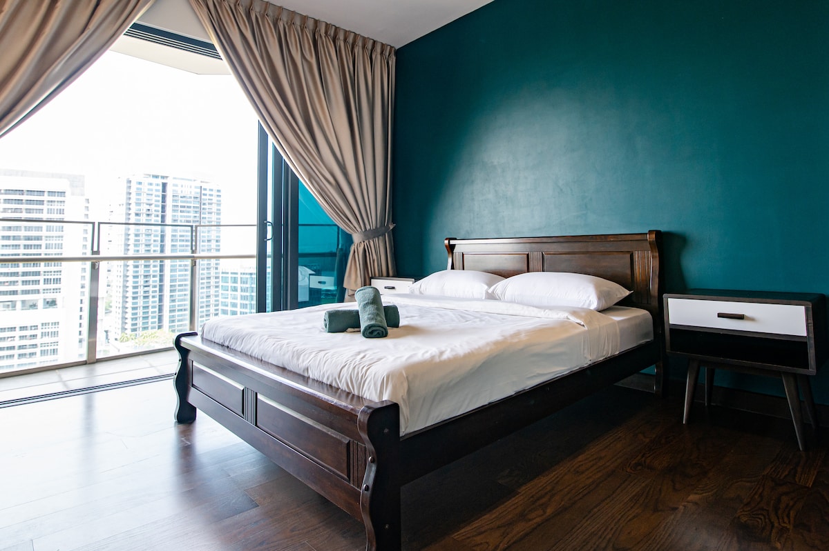 A queen bed with fresh linens and rolled towels is positioned in front of a large window, offering a view of the balconies. The wall is painted a deep blue, while soft drapes frame the window, and a bedside table is placed adjacent to the bed.