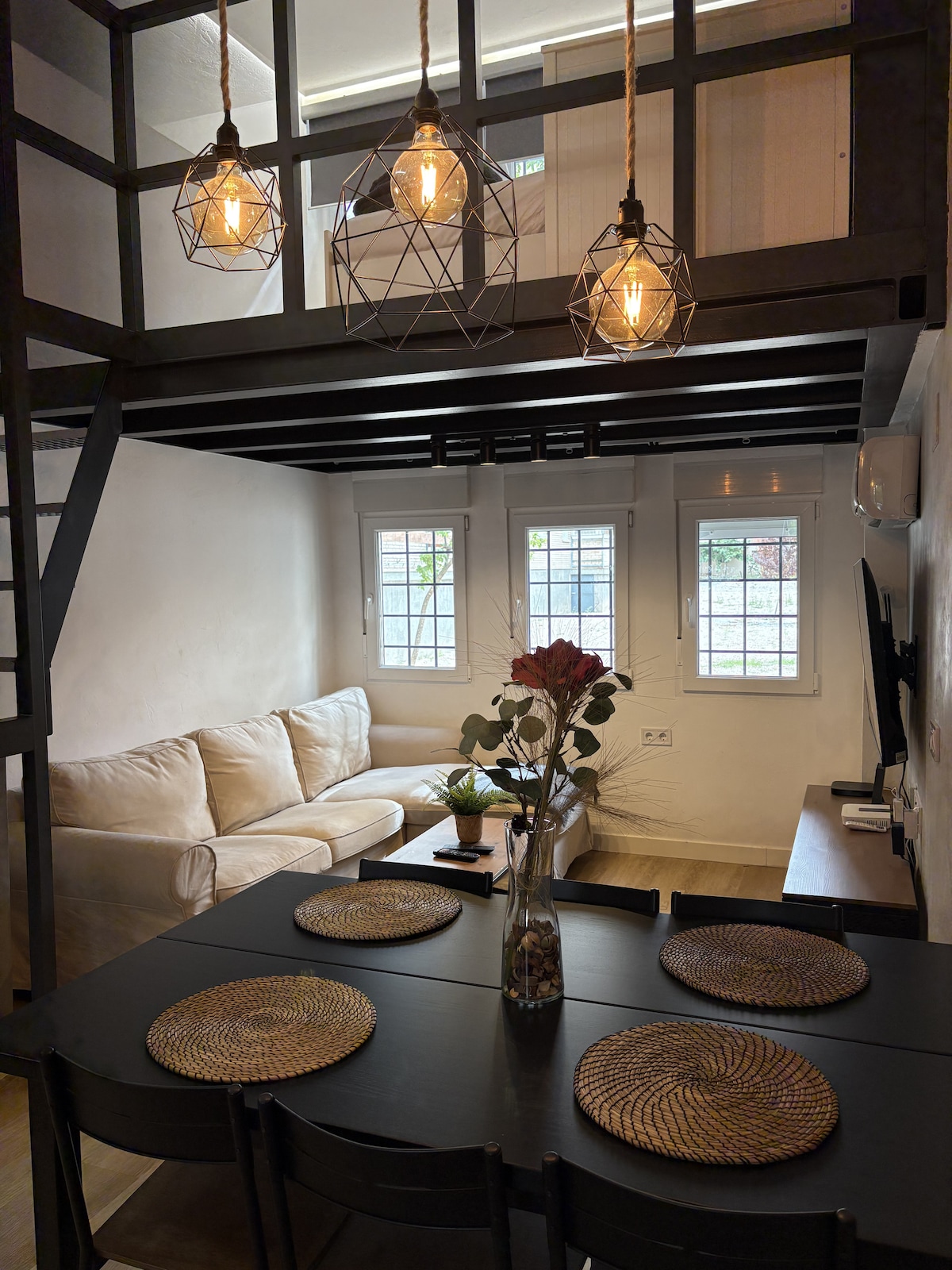 A cozy living area is highlighted by a comfortable couch positioned next to a dining table set for four. Three geometric pendant lights hang from a dark wooden ceiling. Light enters through two windows, and a vase with flowers adds a touch of color.