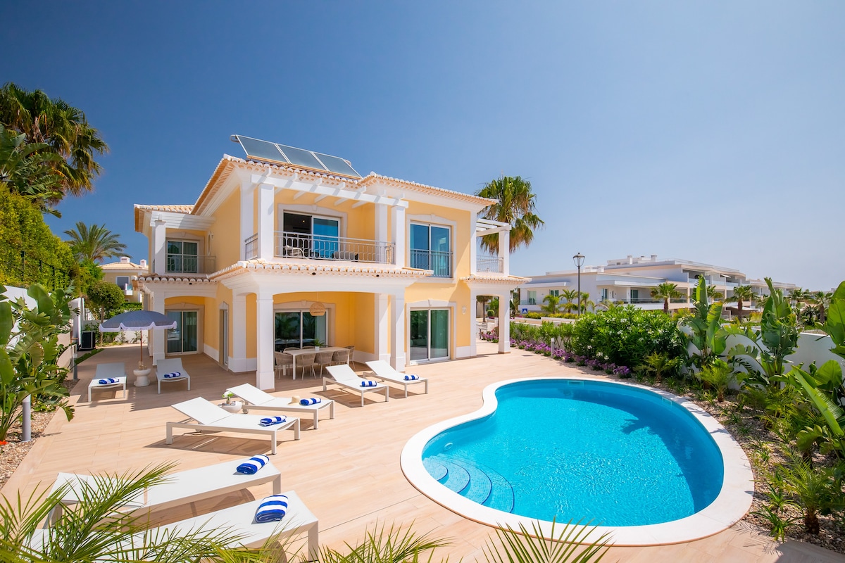 A contemporary villa is set against a clear blue sky, featuring a private curved swimming pool surrounded by sun loungers. Lush green landscaping and tropical plants create a serene outdoor environment, while large glass doors illuminate the inviting interior spaces of the home.