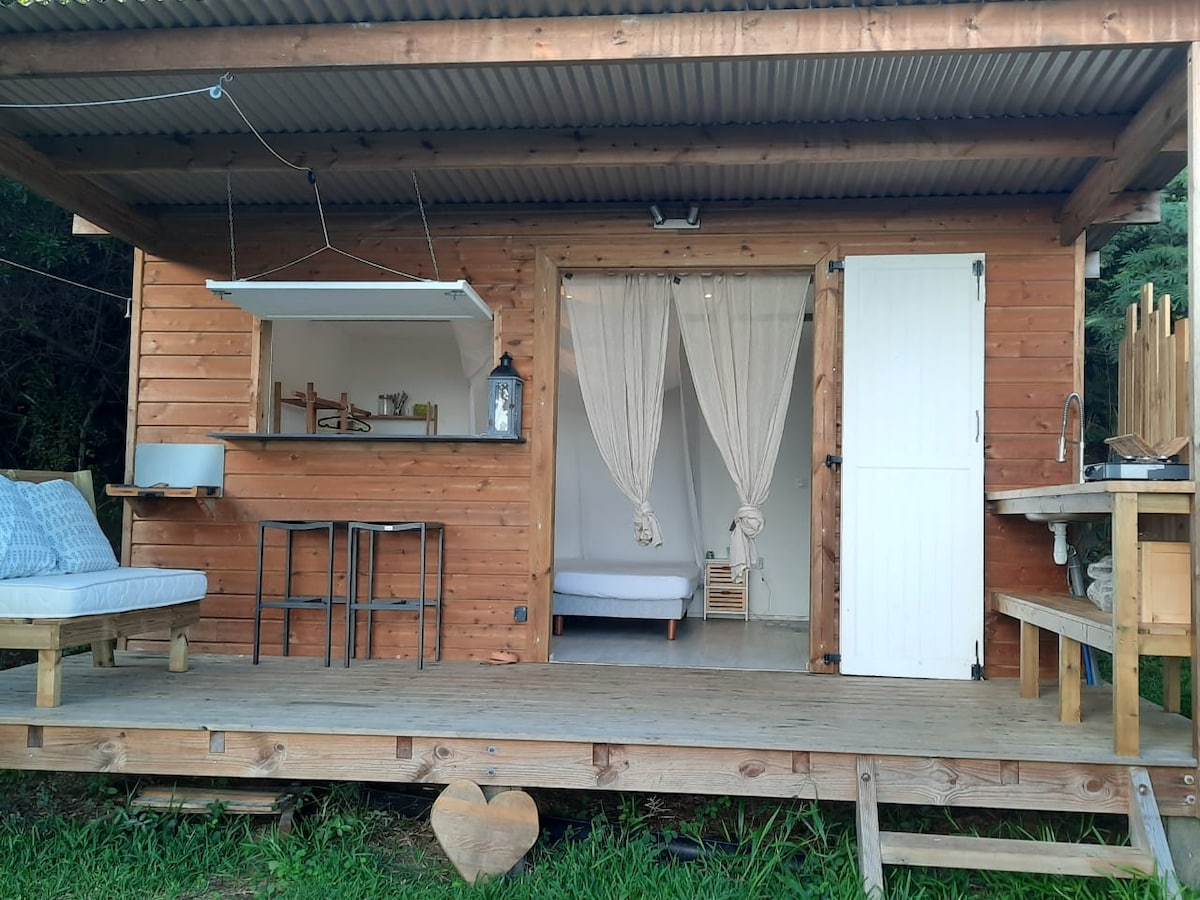 A quaint wooden bungalow is set on a spacious deck, featuring neutral-toned seating and a small dining area. Light-colored curtains drape at the entrance, leading into a simple interior. An outdoor cooking area is equipped with a countertop and functional appliances.