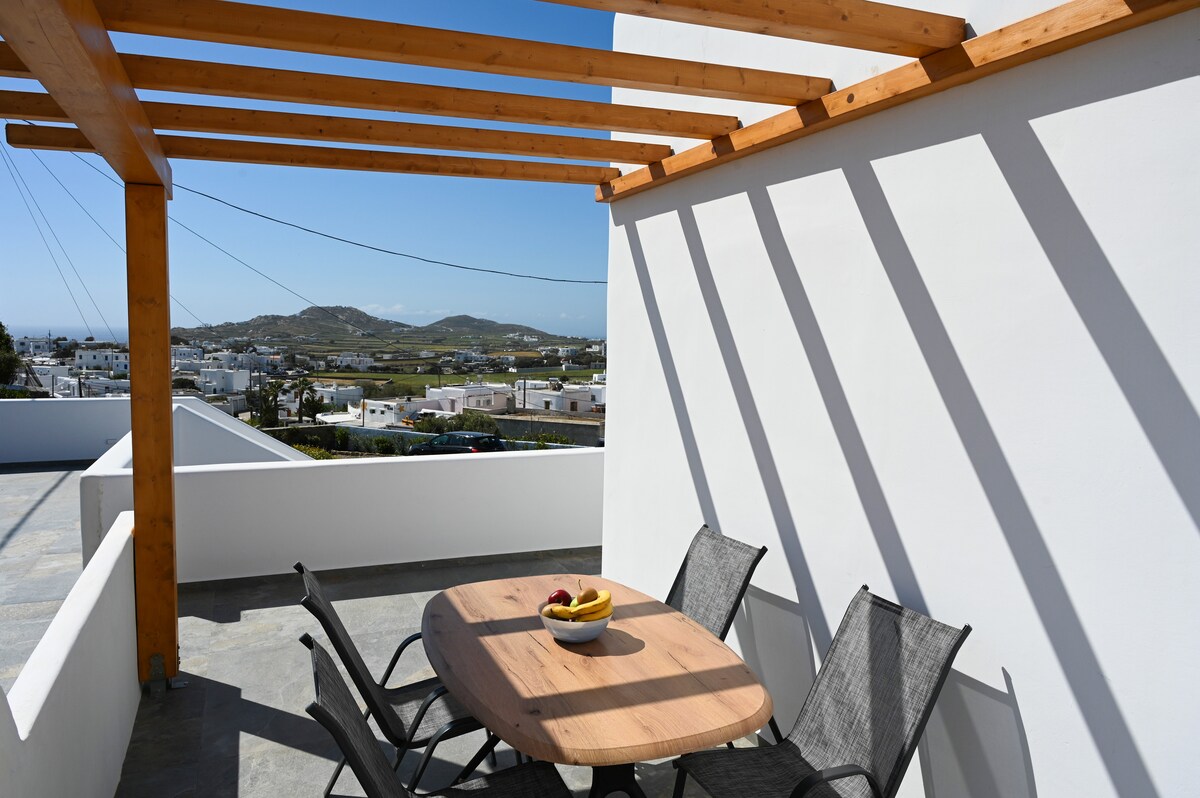 A rooftop terrace is depicted, featuring a circular wooden table surrounded by four textured chairs. Natural light casts shadows on the white walls, while scenic views of distant hills and town houses are visible in the background.