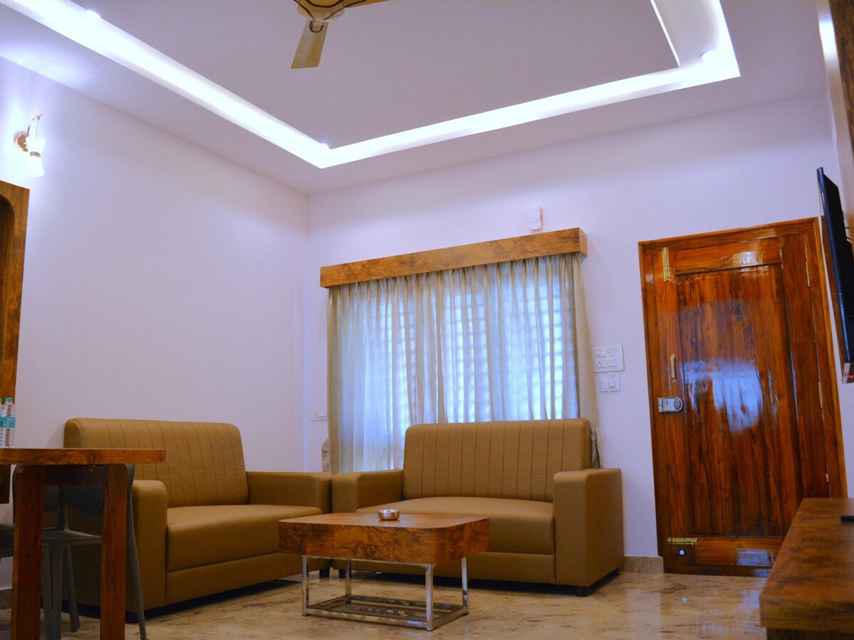 A comfortable living area features two spacious sofas arranged around a glass coffee table. Natural light filters through sheer curtains, illuminating the polished marble flooring. Wooden accents are present in the window and door frames, contributing to the overall warmth of the space.