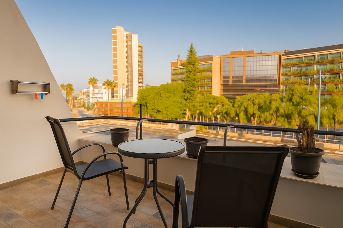 A covered balcony features two black chairs and a round table, surrounded by potted plants. A view of nearby buildings and palm trees is visible in the background, with clear skies enhancing the light atmosphere. The setting offers a relaxing space for outdoor enjoyment.