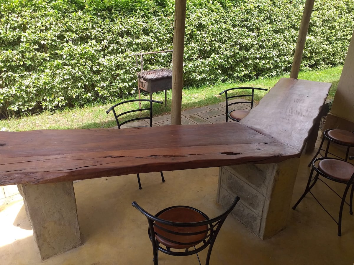 A unique wooden counter table made of natural wood is set on a patio surrounded by greenery. Several black metal chairs are positioned around the table, providing an inviting space for gatherings. A BBQ grill can be seen in the background, nestled against a lush hedge.