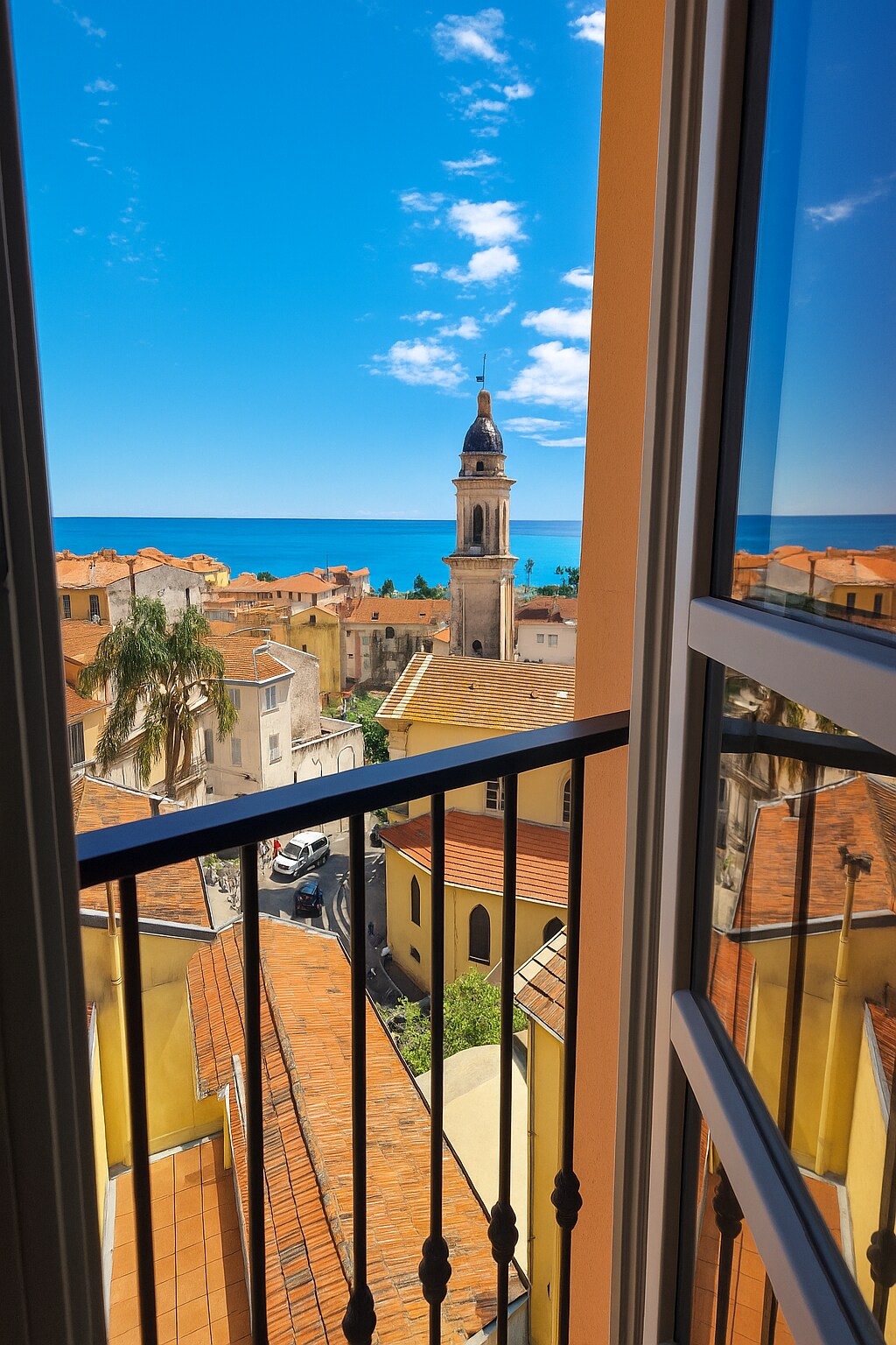 A view from a window showcases a coastal landscape, with a bell tower visible among orange-tiled roofs. The bright blue sky contrasts against the warm colors of the buildings, while the sea can be seen in the distance, enhancing the sense of place.