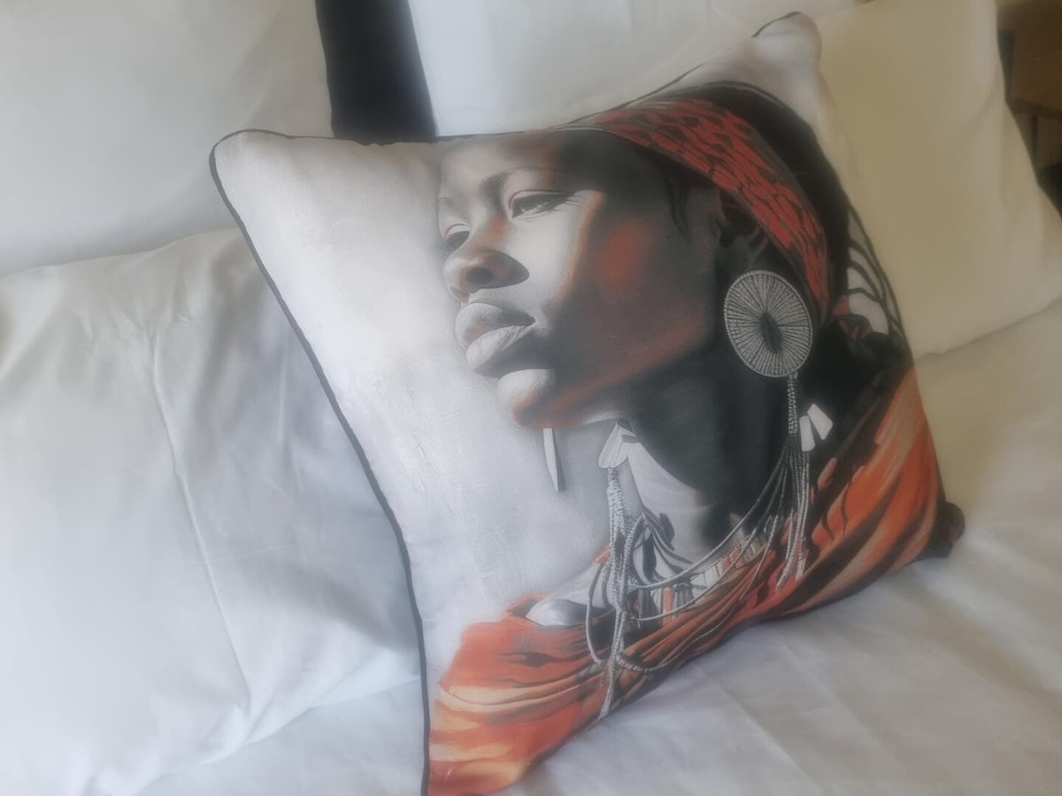 A decorative pillow featuring an artistic design is placed on a spacious bed. The pillow showcases a vibrant image of a figure adorned with traditional accessories, contrasting against the soft white bedding.