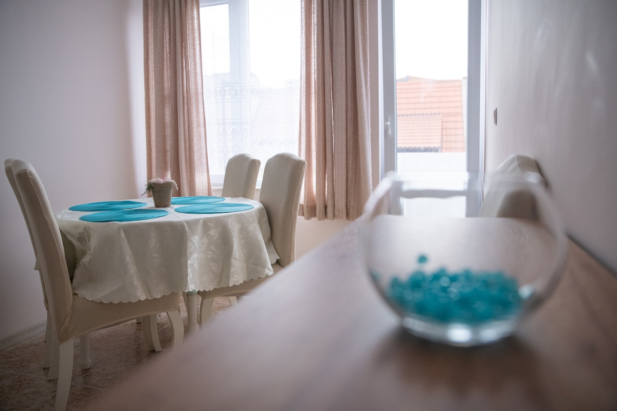 A dining area is presented with a round table set with teal placemats. Four upholstered chairs surround the table. A glass bowl containing blue beads is positioned in the foreground. Light enters through two large windows, partially covered by sheer curtains.