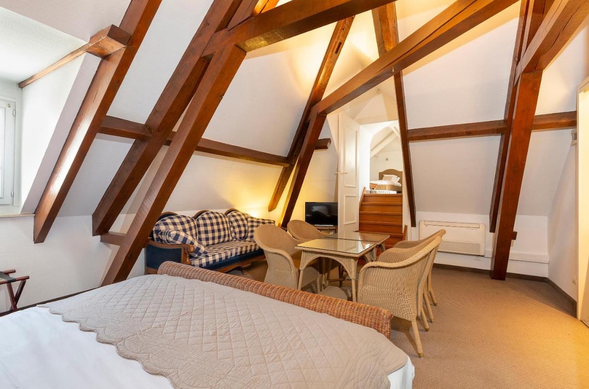 A cozy attic space is featured, showcasing sloped wooden ceilings and exposed beams. A spacious bed is accompanied by a seating area with a checkered sofa and glass coffee table. Light is provided by large windows, creating a bright and inviting atmosphere.