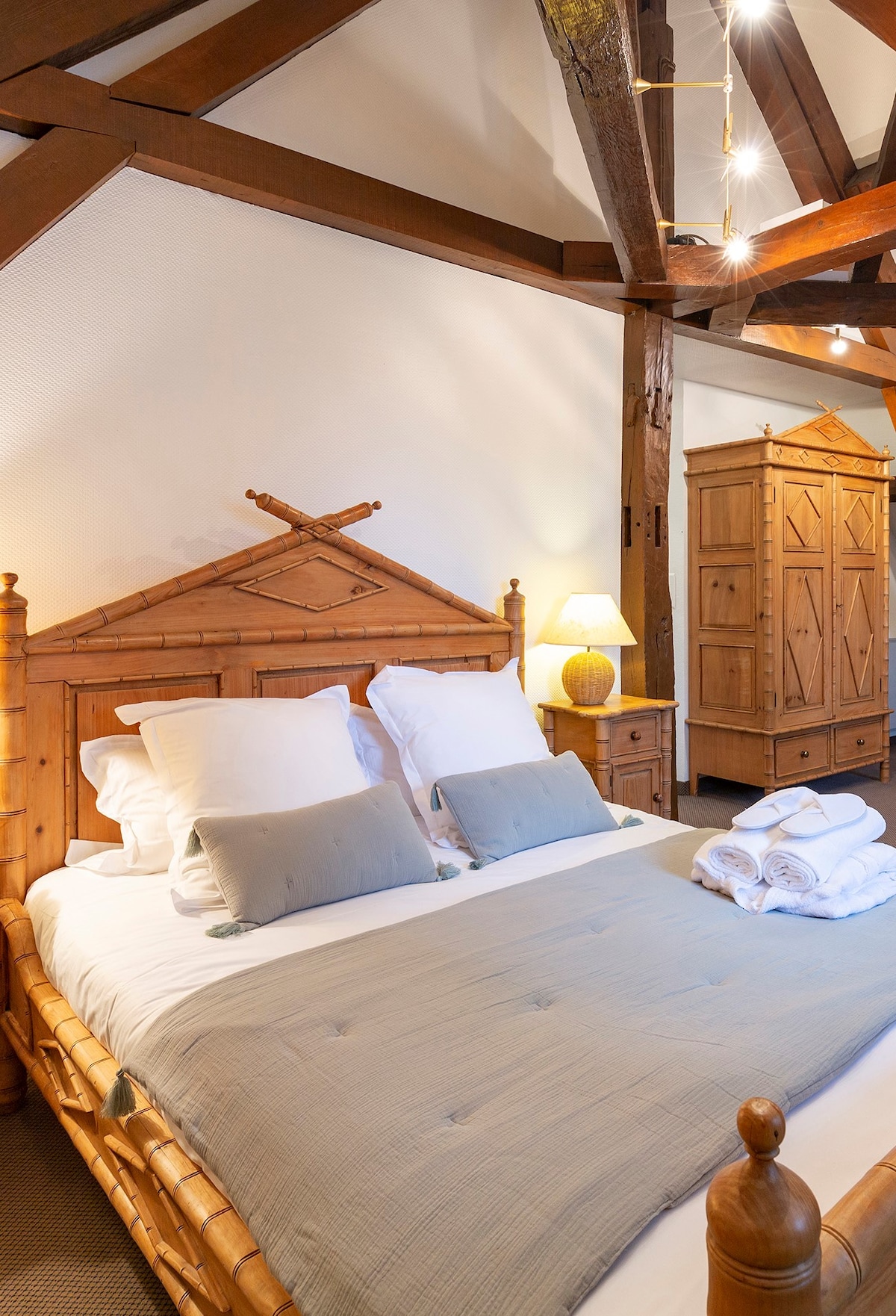 A spacious bedroom features a wooden bed frame with a decorative headboard. The bed is adorned with multiple pillows and a light-colored quilt. A bedside lamp provides soft lighting, while a cabinet with intricate design elements stands in the background.