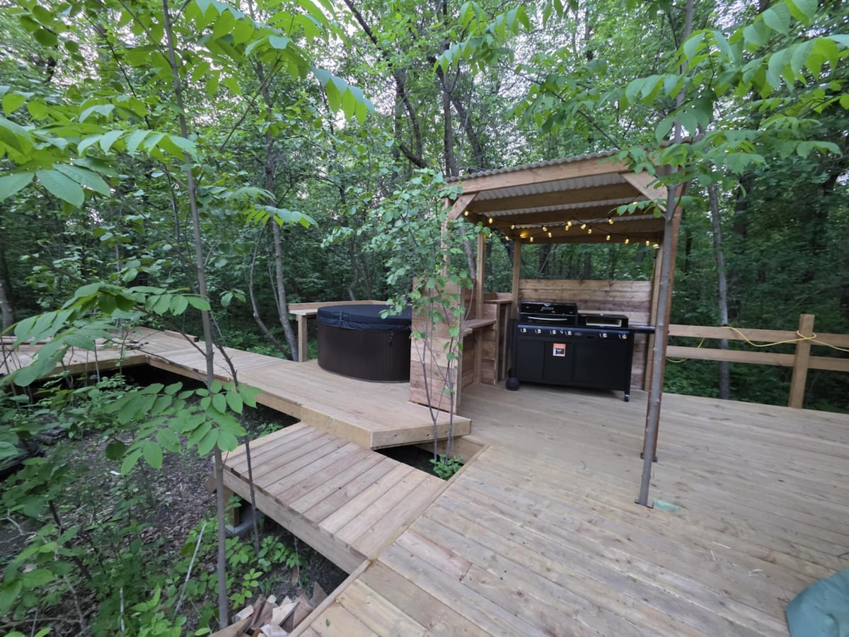 An outdoor area is set among trees, featuring a wooden deck with soft lighting. A hot tub is positioned opposite an outdoor cooking area that includes a grill. The surroundings depict a natural, serene environment for relaxation.
