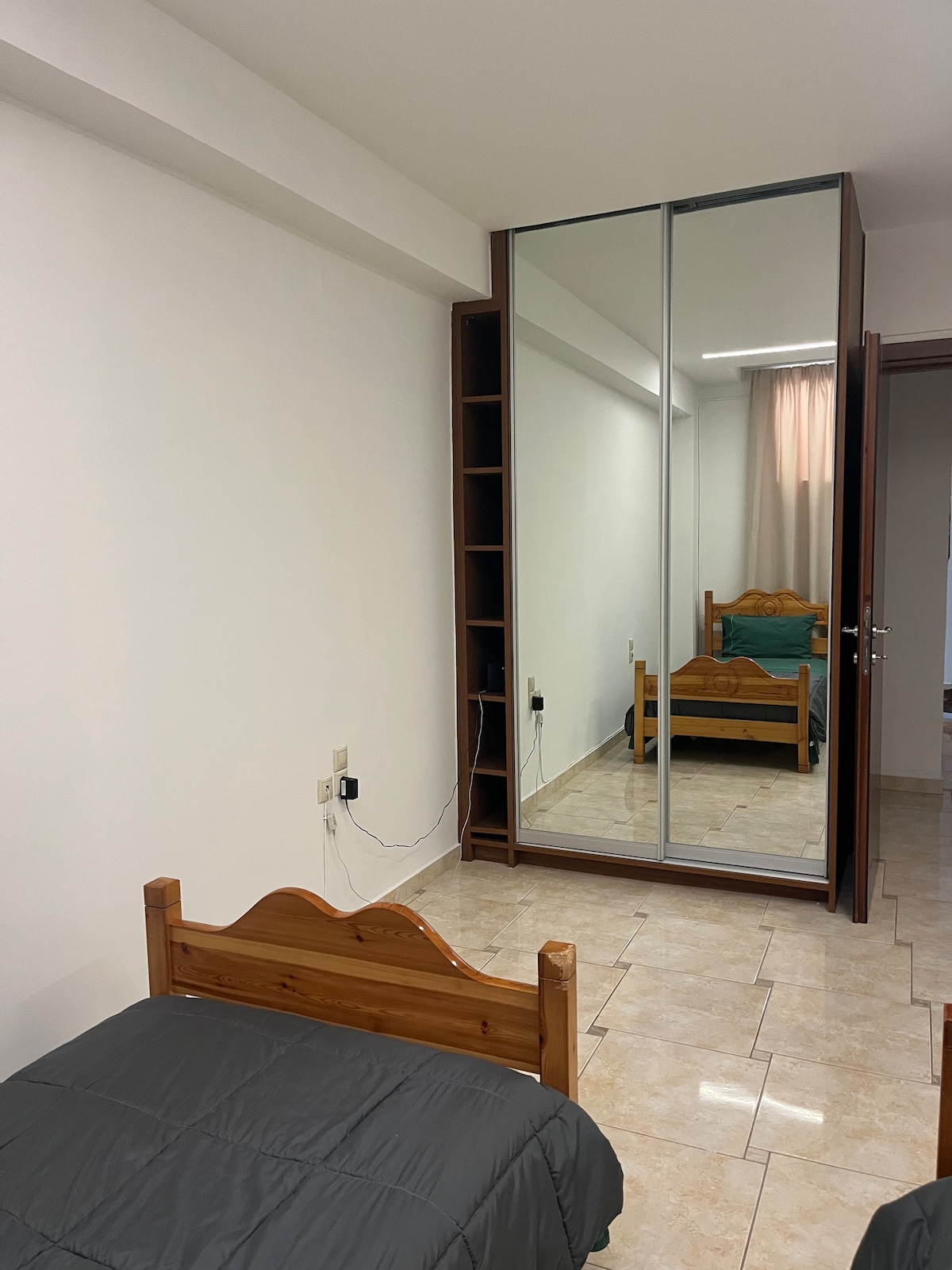 A well-lit bedroom features a wooden single bed with a dark blanket. Beside the bed, a full-length mirror is mounted on a large wardrobe. A soft green bedspread is visible on the adjoining bed, which is positioned against a light-coloured wall.