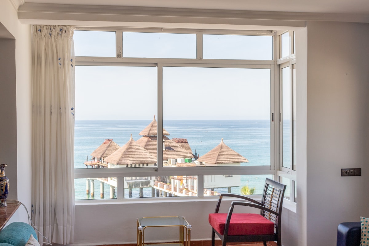 A large window frames a clear view of the ocean, showcasing calm blue waters and a distinctive restaurant with a thatched roof. A wooden chair with red upholstery and a glass table are positioned in front of the window, creating a comfortable seating area.