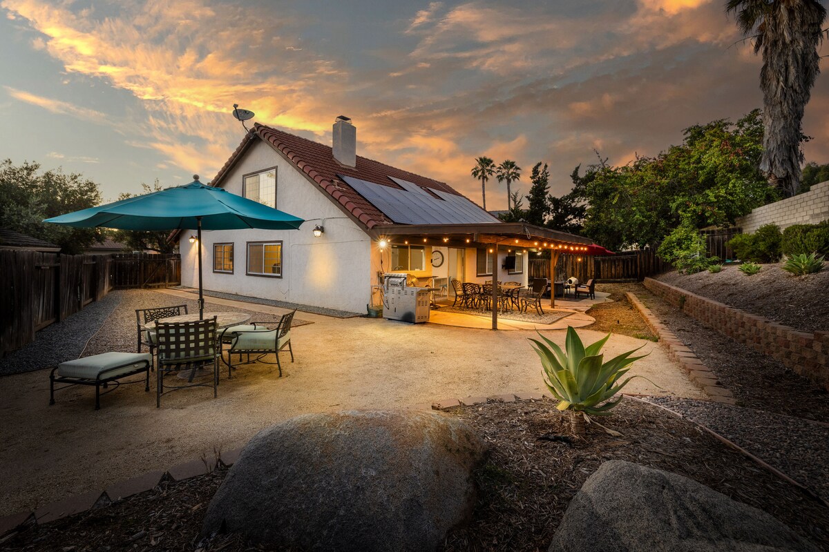 An inviting outdoor space is presented, featuring a patio with seating under a large umbrella. The fenced backyard includes decorative stones and landscaping elements. The home is illuminated by warm lights, showcasing large windows and a spacious patio area, ideal for gatherings.