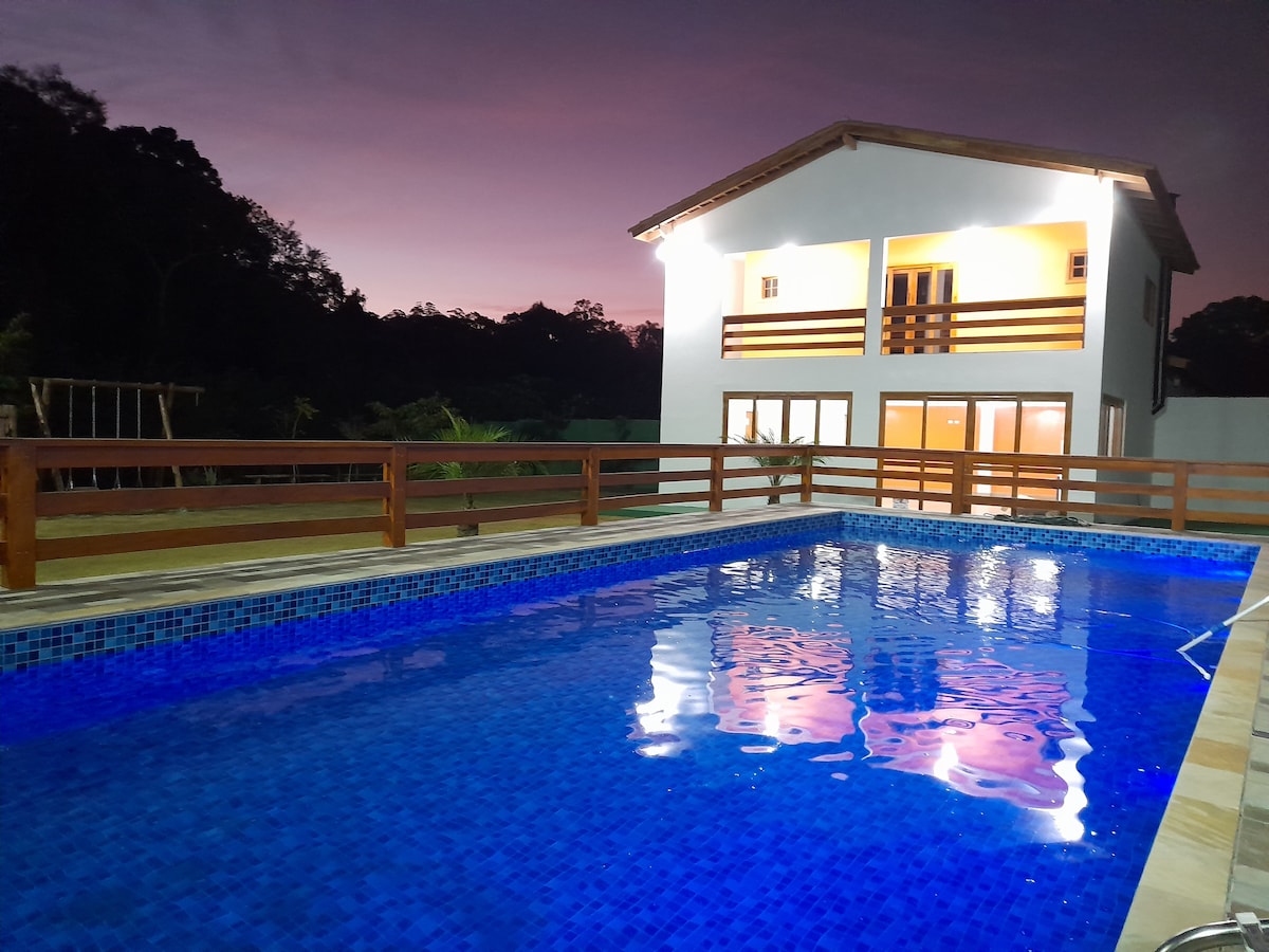 A spacious swimming pool is centered in the foreground, its bright blue water reflecting the evening sky. The two-story house in the background features large windows and wooden railings on the balcony, surrounded by greenery and soft lighting.