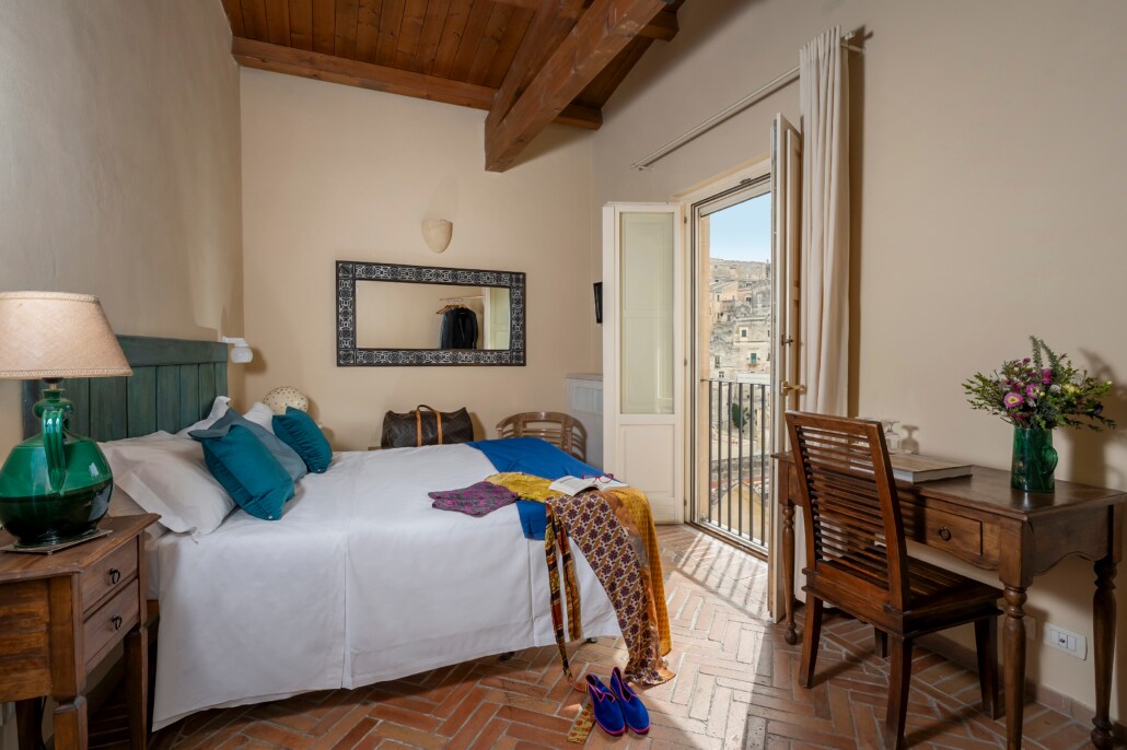 A spacious room features a large bed adorned with white linens and blue accent pillows. A wooden desk and chairs are positioned nearby, with a vase of flowers adding a touch of color. A balcony door opens to reveal views of the surrounding Sassi di Matera.