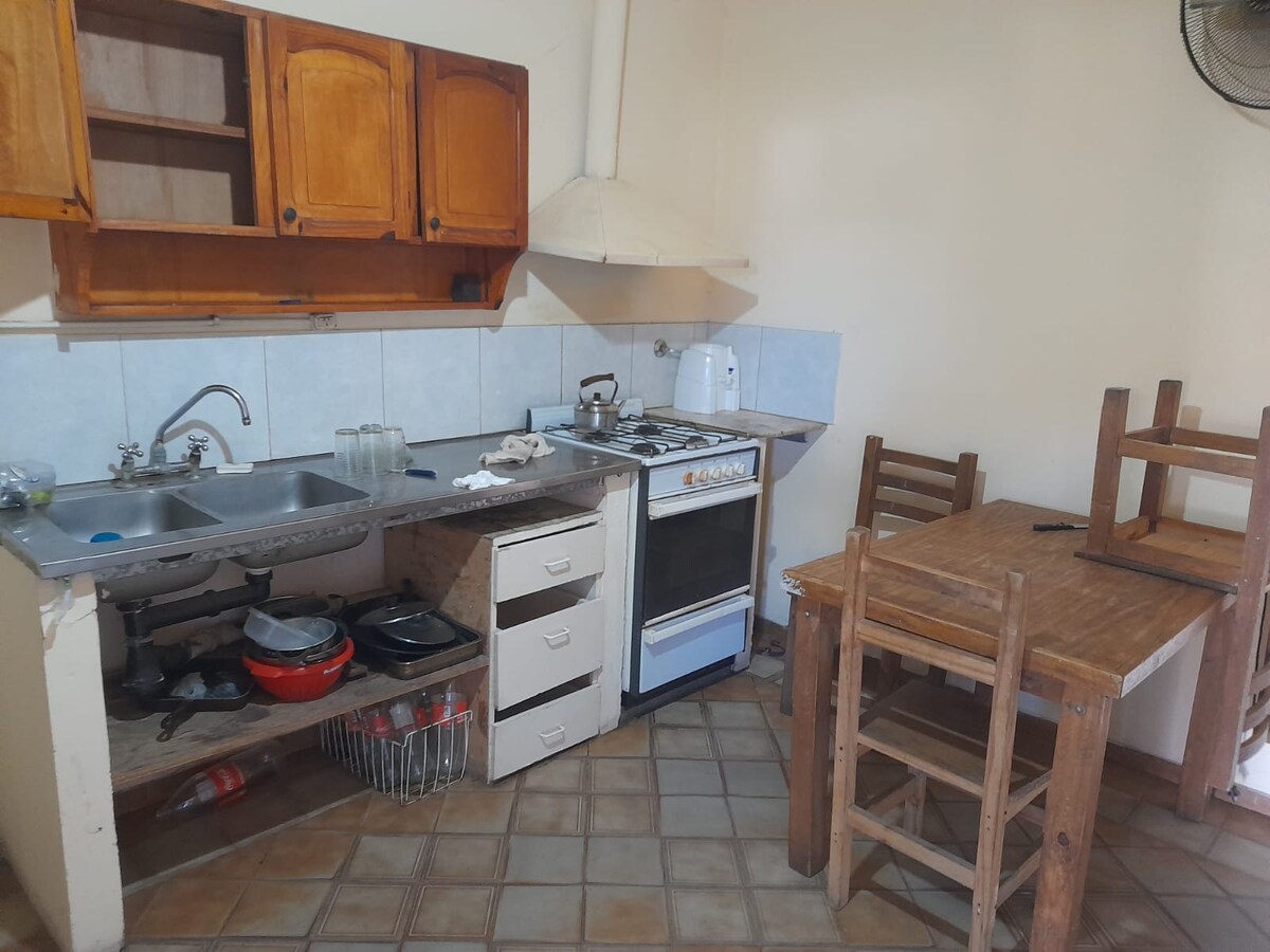 A functional kitchen area features wooden cabinets and a gas stove. Stainless steel sink and countertop are equipped with basic kitchenware. A wooden dining table with three chairs stands nearby, providing a simple dining space. Tile flooring adds practicality to the area.