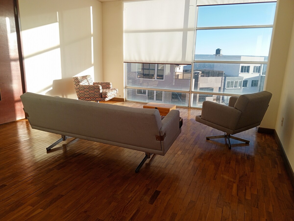 A bright living area is filled with natural light, featuring a modern gray sofa and a patterned armchair. A wooden coffee table is positioned between the two seating arrangements. Large windows offer a view of the surrounding cityscape.