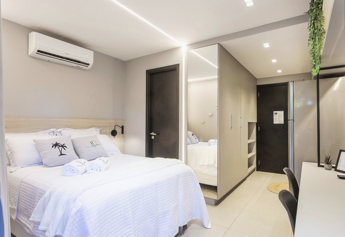 A modern bedroom features a spacious bed with white bedding and decorative pillows. A sleek wardrobe is visible, along with a desk and chair. Natural light is provided by overhead lighting, and an air-conditioning unit is mounted on the wall.