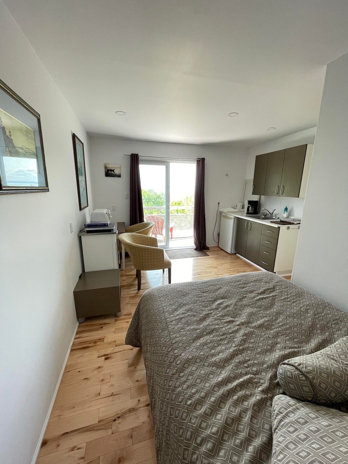 A compact mini-studio features a spacious bed with a textured blanket. A pair of chairs are positioned near a kitchenette, which includes a small fridge and coffee maker. Natural light fills the room through sliding glass doors that lead to a private balcony overlooking greenery.
