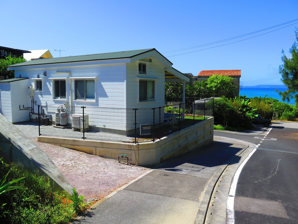 A small log cabin-style house is presented, featuring a covered terrace and enclosed yard. The property is set against a clear blue sky, with the calming ocean visible in the background. Nearby greenery and the structure's white facade contribute to a serene coastal environment.