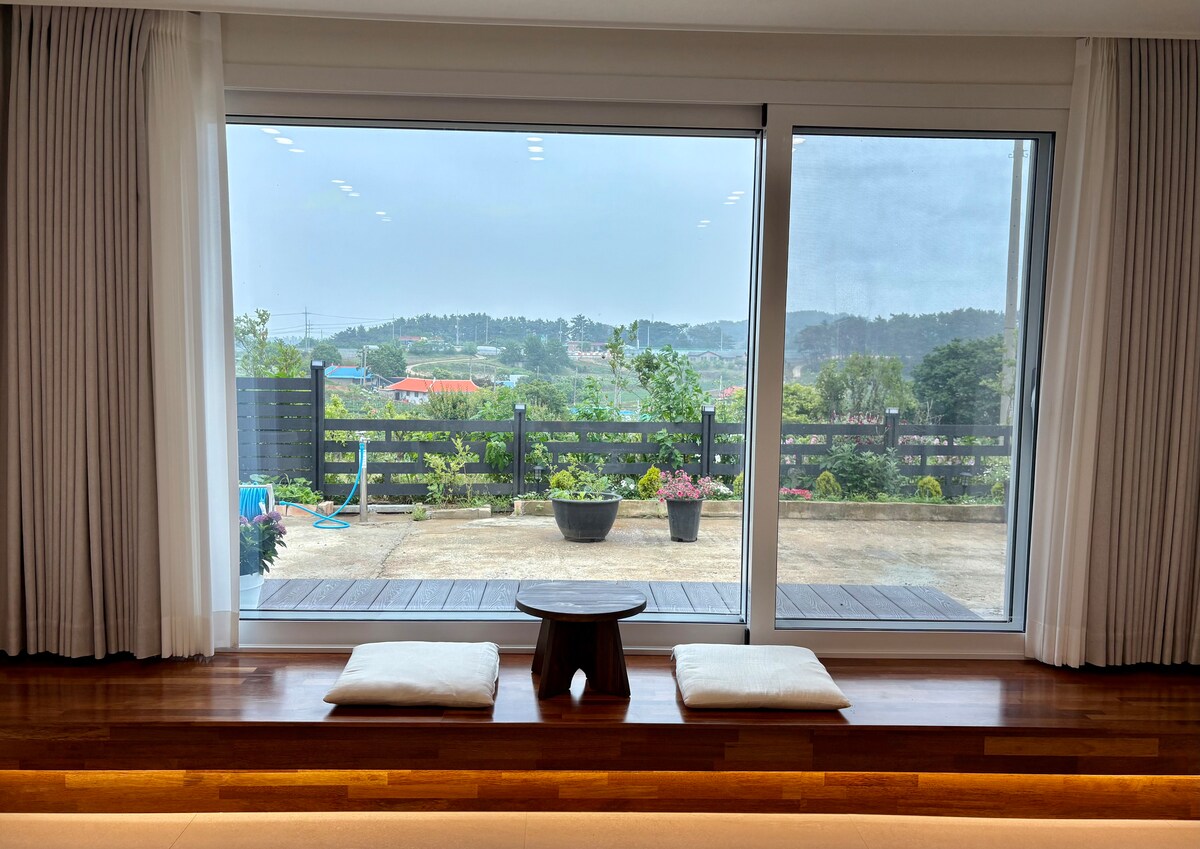 A large window offers a view of the outdoor area, surrounded by greenery and a well-maintained garden. Two floor cushions are placed on a wooden ledge, providing a cozy spot to enjoy the scenery. The space is brightly lit with natural light.