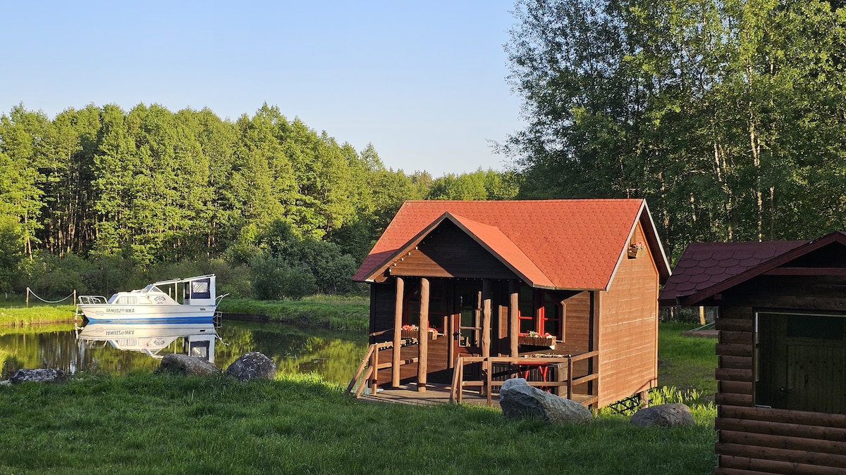 A wooden cabin with a red roof is situated beside a tranquil pond, surrounded by lush greenery. A small boat is moored nearby, and picnic-style seating is arranged on the cabin's deck, providing an inviting spot to enjoy the natural landscape.