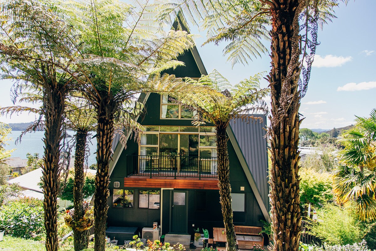 A distinctive A-frame house is framed by tall ferns, showcasing expansive windows that provide views of the surrounding landscape. A wooden deck extends from the front, with a welcoming outdoor seating area. Lush greenery surrounds the property, adding to the tranquil setting.