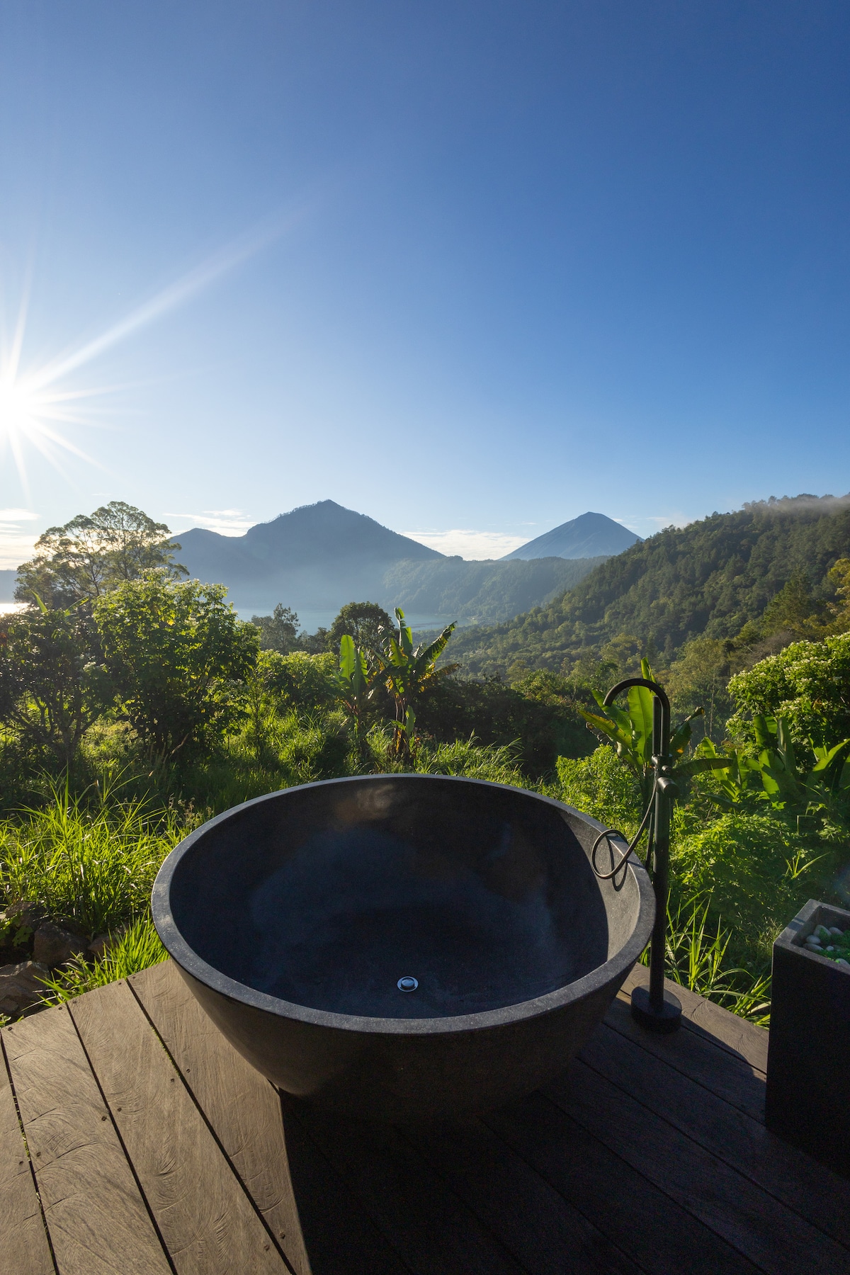 An outdoor stone bathtub is positioned on a wooden deck, providing an unobstructed view of distant mountains and lush greenery. The sky reflects morning light, enhancing the serene atmosphere. The design offers an opportunity for relaxation amidst natural beauty.