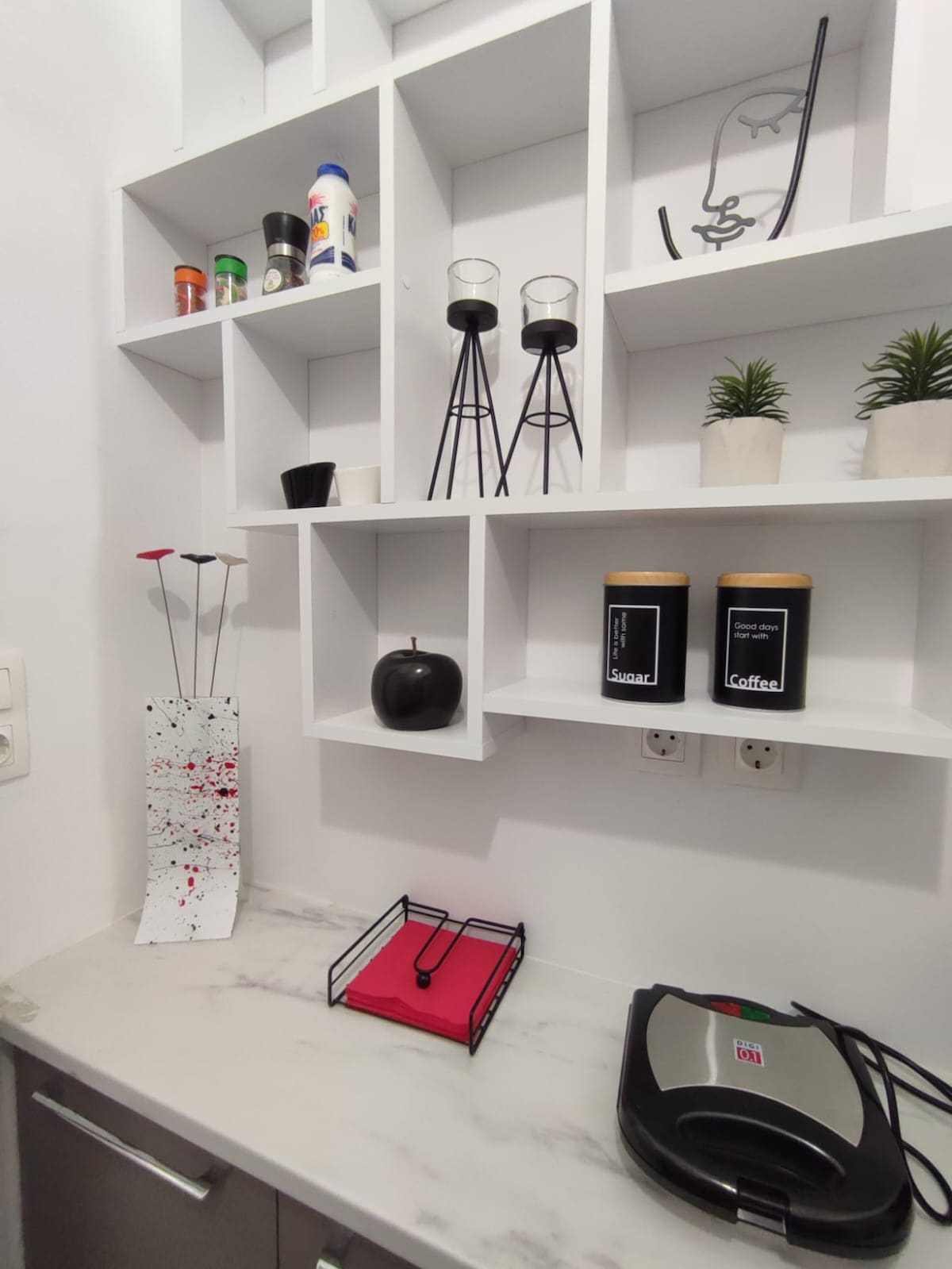 A modern kitchen corner is presented, featuring white shelving that displays spices, decorative items, and small potted plants. A marble countertop holds a coffee maker and a black storage container, while a vibrant, artistic vase with red accents adds a touch of color.