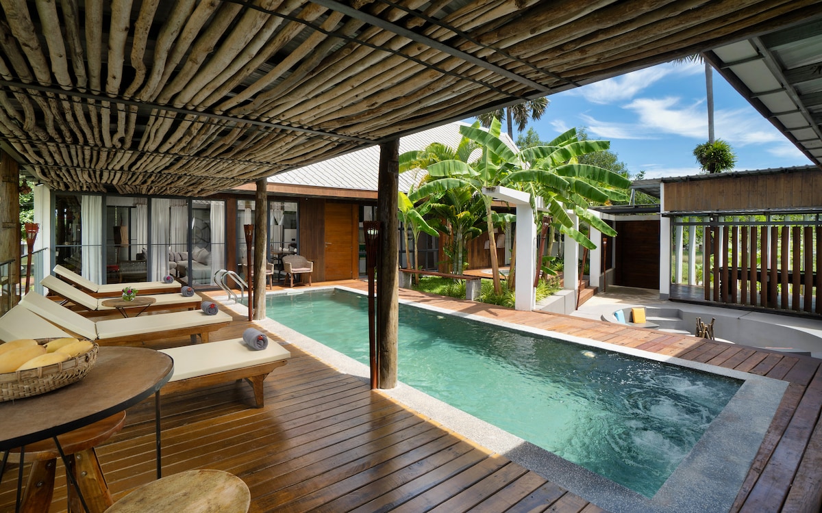 An inviting outdoor area features a private pool surrounded by sun loungers atop a wooden deck. Lush greenery and tropical plants create a serene backdrop, while the thatched roof of the structure offers shade to the seating space nearby.