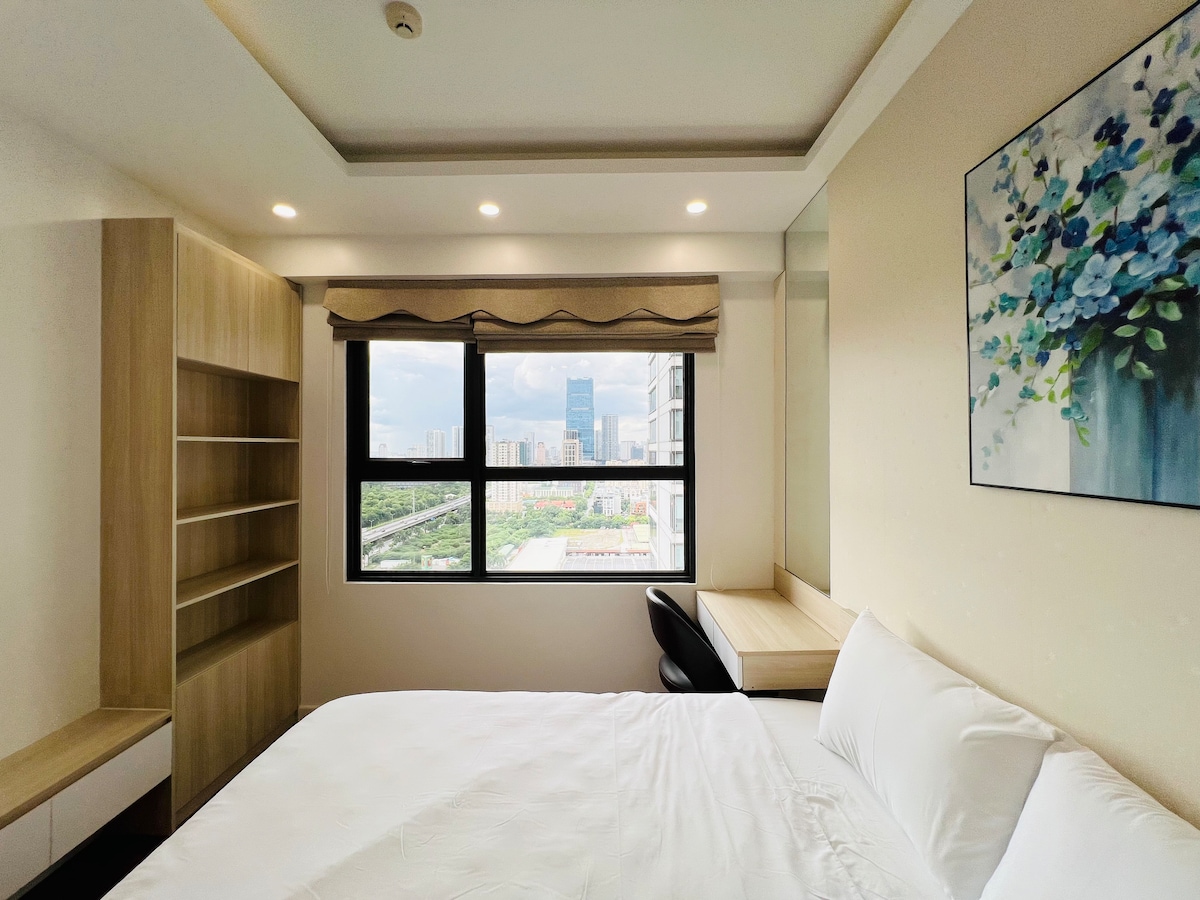 A well-lit bedroom showcases a spacious bed with crisp white linens. A large window offers a view of the city, while natural light fills the room. Nearby, a desk and chair are positioned next to a wooden shelving unit, adding functionality to the space.