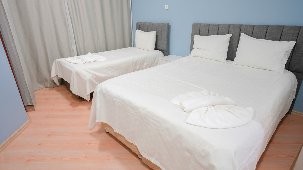 The sleeping area features two beds, one spacious and one smaller, both dressed in white linens. Gray headboards contribute to the modern design. Light wooden flooring contrasts with the soft colors of the walls, while sheer curtains frame a window, allowing natural light into the room.