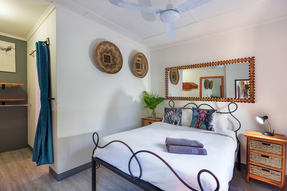 A spacious bed with a decorative metal frame is positioned against a light-colored wall. Textiles in various shades are arranged, including two rolled towels. Natural elements are introduced through a potted plant and woven wall hangings, complemented by a large mirror reflecting the room's light.