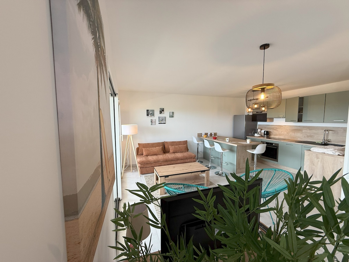 An open living space is featured, with a comfortable sofa facing a modern kitchen equipped with sleek appliances. Dining chairs surround a wooden table, and decorative wall art enhances the atmosphere. Green plants add freshness, while large windows allow natural light to brighten the room.