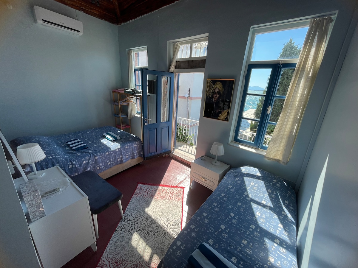 A well-lit guest room features two beds with patterned bedding, a small table, and a white nightstand. Large windows allow natural light to fill the space, while a blue door leads to a balcony offering views of the surroundings.
