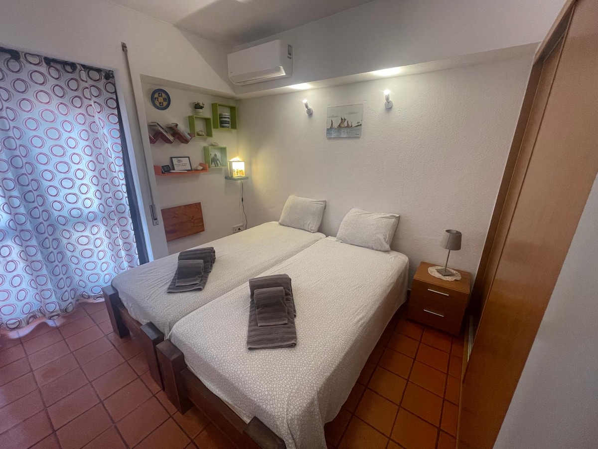 The sleeping area features two spacious beds with light-colored bedding and neatly arranged towels placed on top. A bedside table with a lamp is positioned beside one bed, and a small shelf with decor items is visible against the wall. Natural light enters through a window covered with patterned curtains.