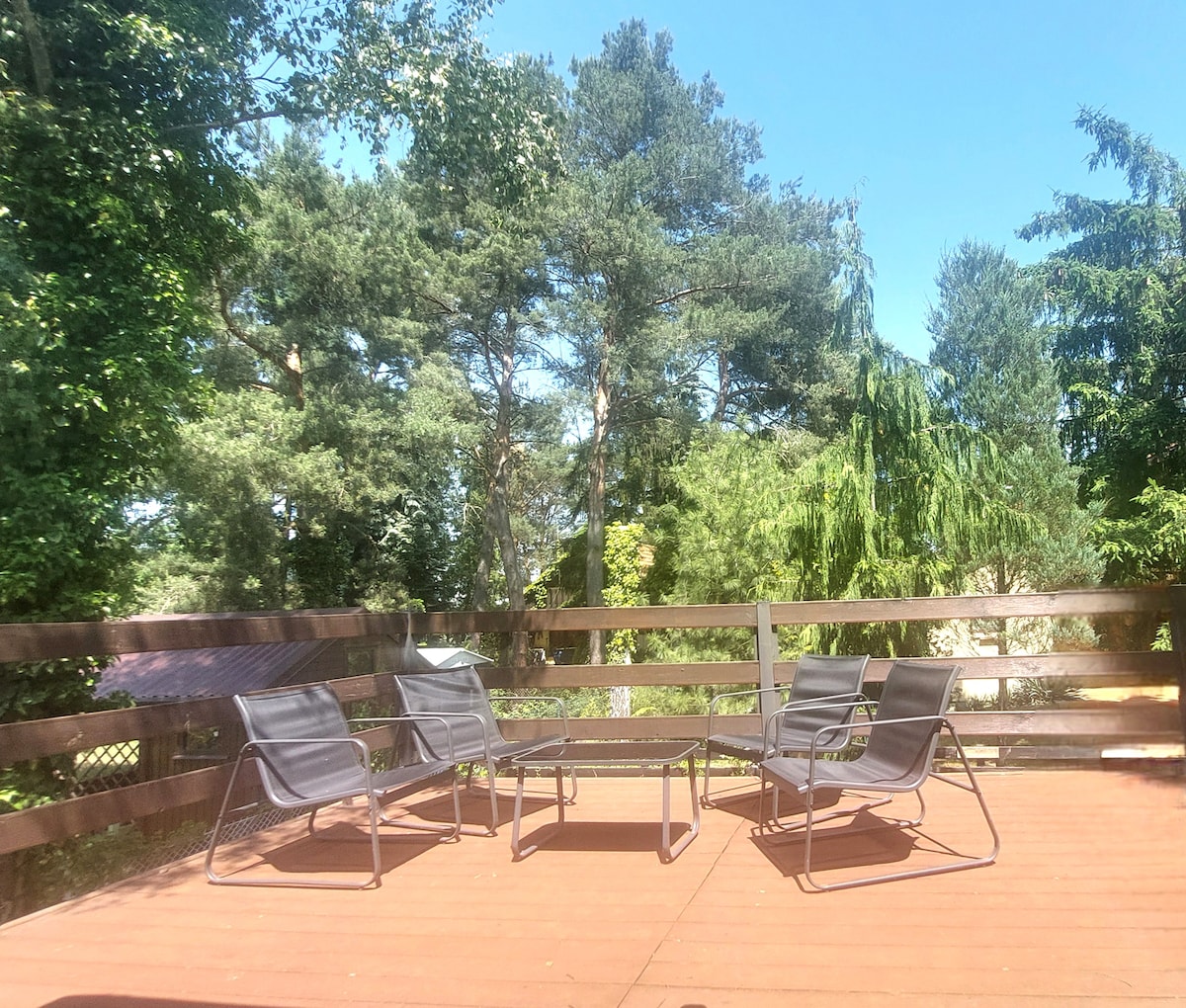 A wooden terrace features a set of four black lounge chairs arranged around a small table. Surrounding trees provide a natural backdrop, enhancing the sense of privacy and tranquility in the outdoor space.