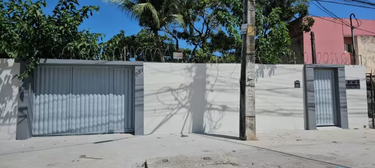 The exterior of the property is presented, featuring a light-colored wall surrounded by greenery. A large gate is visible on the left side, while shadows from tree branches are cast on the wall. A utility pole stands on the right, contributing to the urban setting.