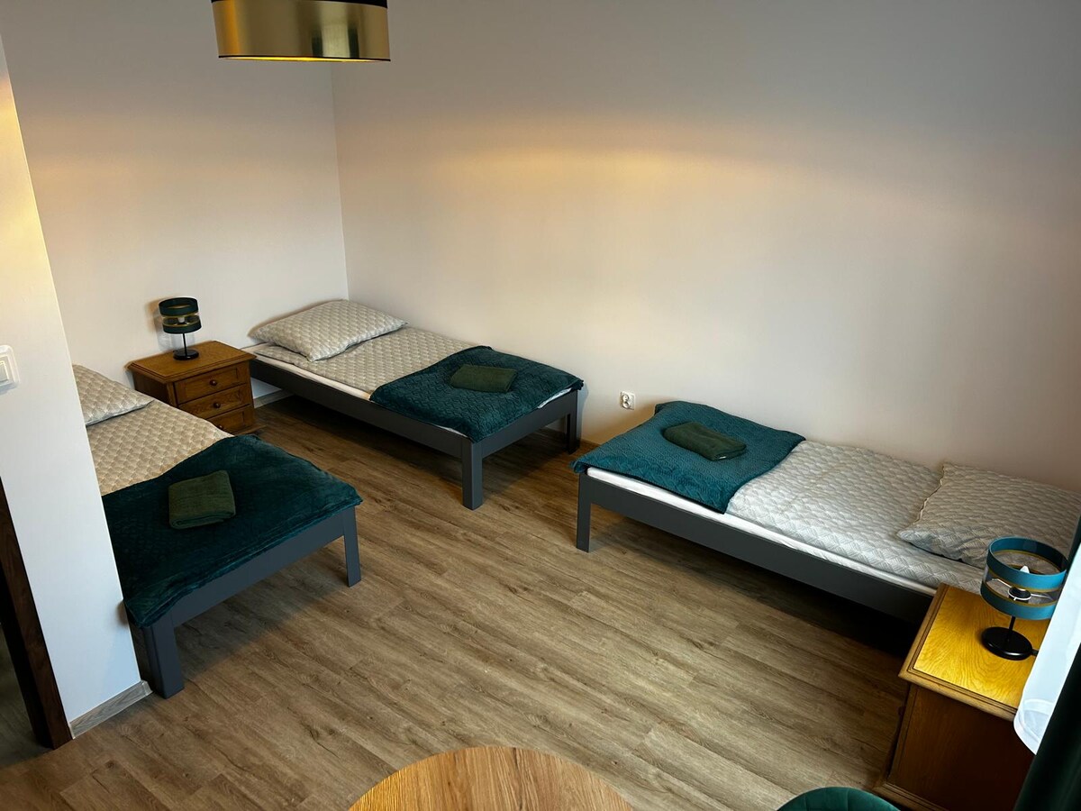 The room features three single beds neatly arranged, each topped with soft blankets and towels. A wooden nightstand is positioned beside one bed, while a table lamp adds a gentle illumination. The natural wood flooring contributes to the space's cozy ambiance.