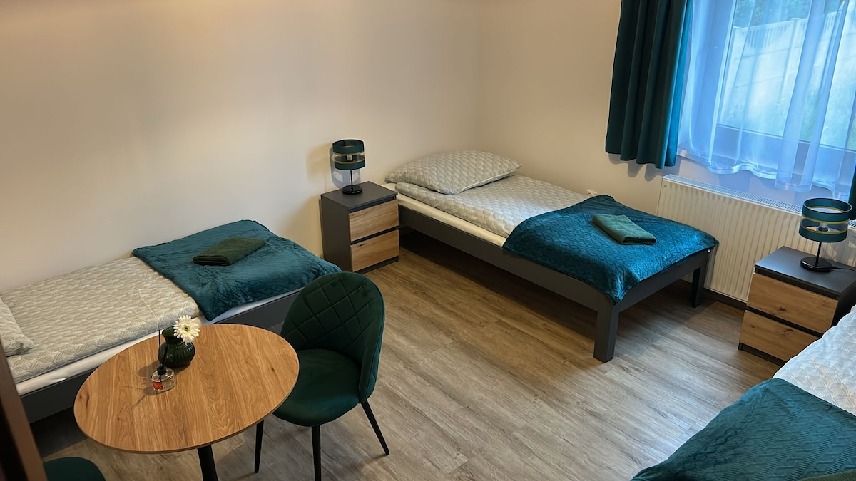 A well-lit bedroom is equipped with three individual beds, each adorned with green linens. A small table with two chairs occupies the center of the room, while a pair of nightstands with lamps stands beside the beds. Natural light filters through the sheer window curtains.