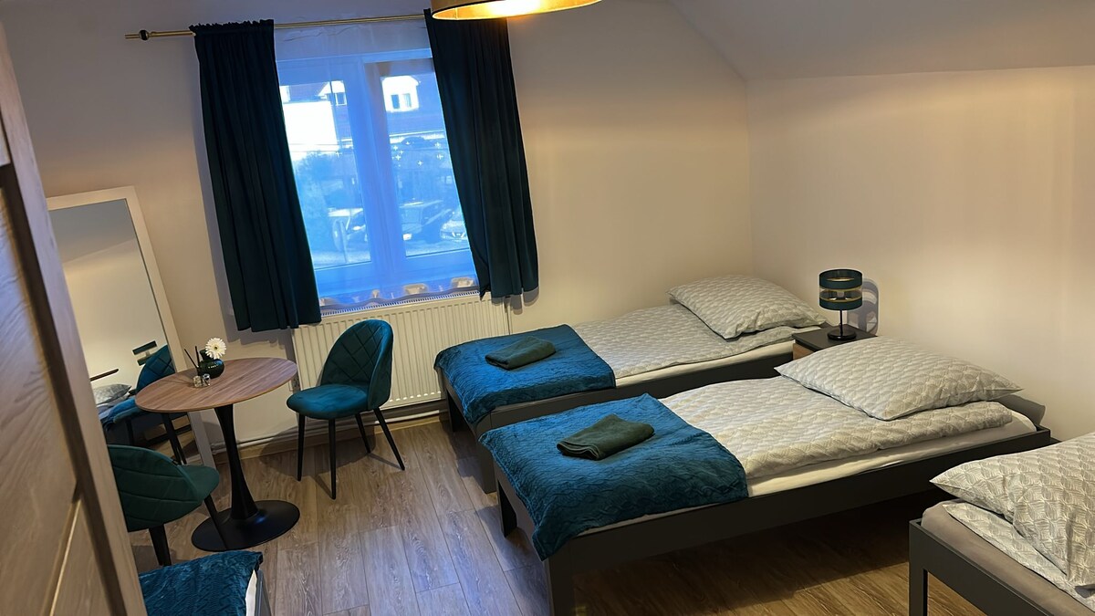 A welcoming bedroom features two spacious beds with teal accents, complemented by matching towels. A small round table is paired with two chairs, situated near a large window that allows natural light to fill the room. Soft lighting is provided by an overhead lamp.
