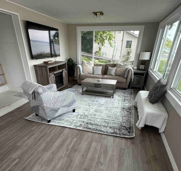 A cozy living area features a comfortable sofa and armchair, positioned on a soft rug. A large window offers a view of the outdoors, while a 75" Smart TV is mounted on the wall. Natural light brightens the space, enhancing its inviting design.