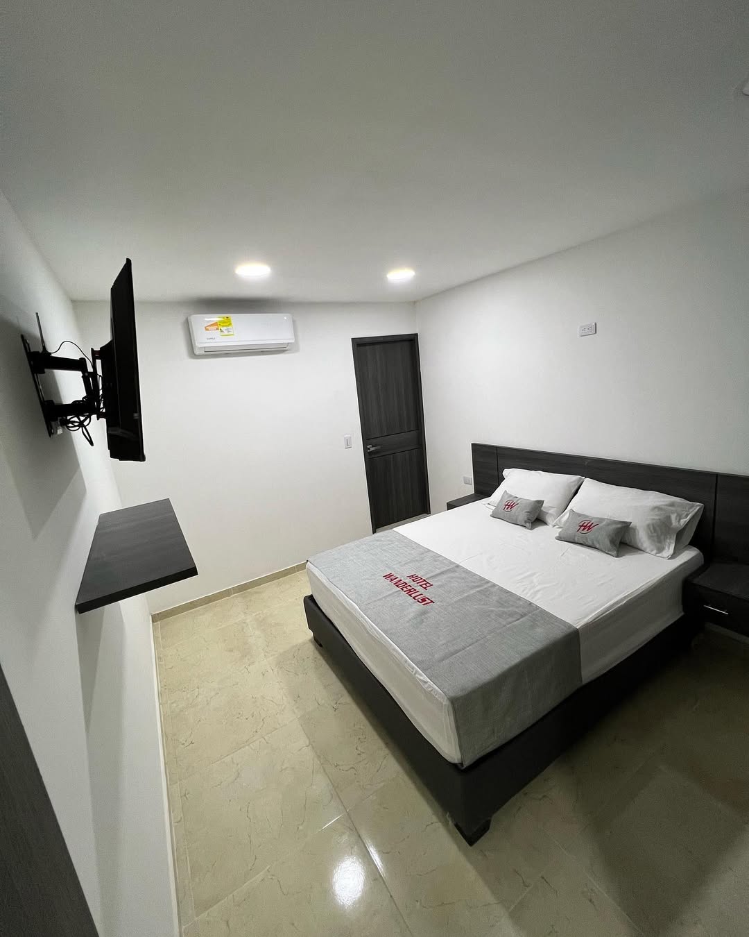 The well-lit bedroom features a spacious bed with a light grey and white color scheme. A television is mounted on the wall, and minimalistic shelving provides additional storage. The room is designed with sleek, modern furnishings against a neutral backdrop, contributing to a clean and calm environment.