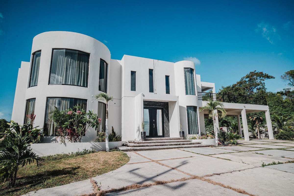 A modern, elegantly designed exterior of a white building is visible, featuring large windows that allow natural light to enter. Lush greenery surrounds the entrance, accented by tropical plants. The smooth, clean lines of the architecture create a striking visual against the blue sky.
