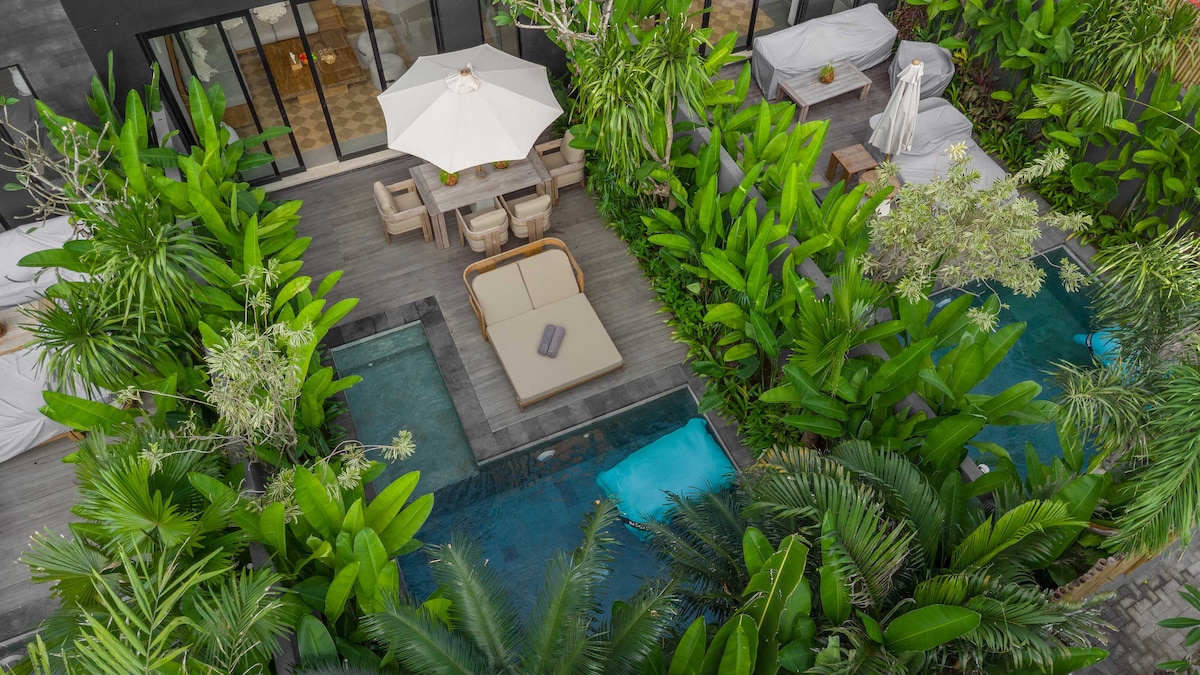An aerial view showcases a private pool area surrounded by lush greenery. Two rectangular pools are located beside a sun loungers area, complete with an umbrella. Comfortable seating is visible nearby, nestled amid tropical plants providing an intimate atmosphere.